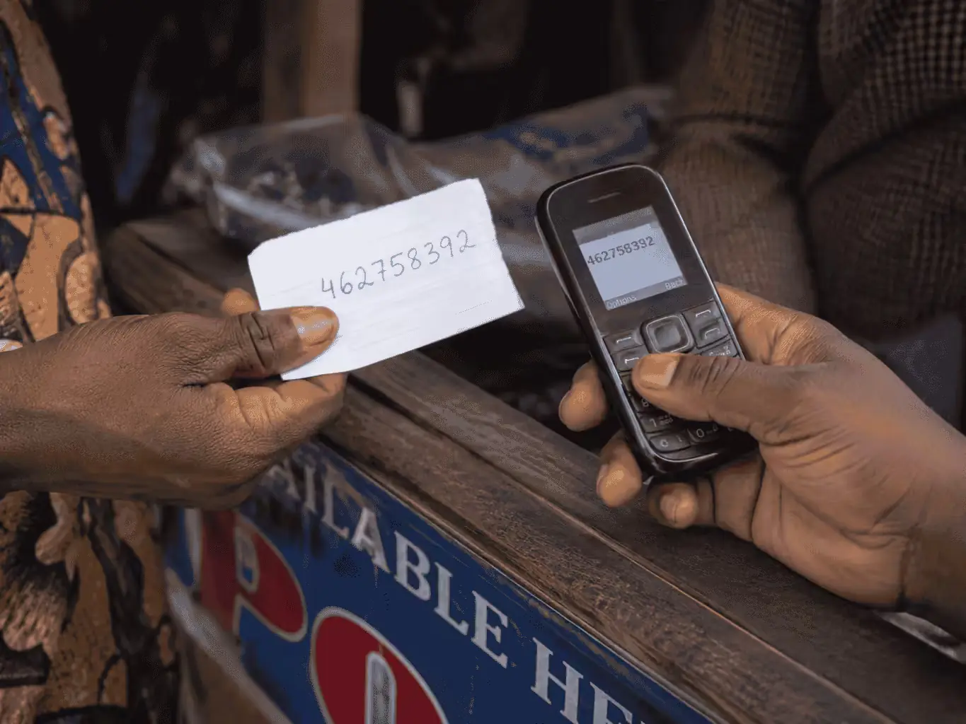 A handwritten 9-digit Ticket IT code on a paper slip, being keyed into a basic feature phone at a stall
